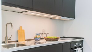 A modern kitchen with dark cabinets, a stainless steel sink, a wooden cutting board, a patterned pitcher, and a bowl of apples.