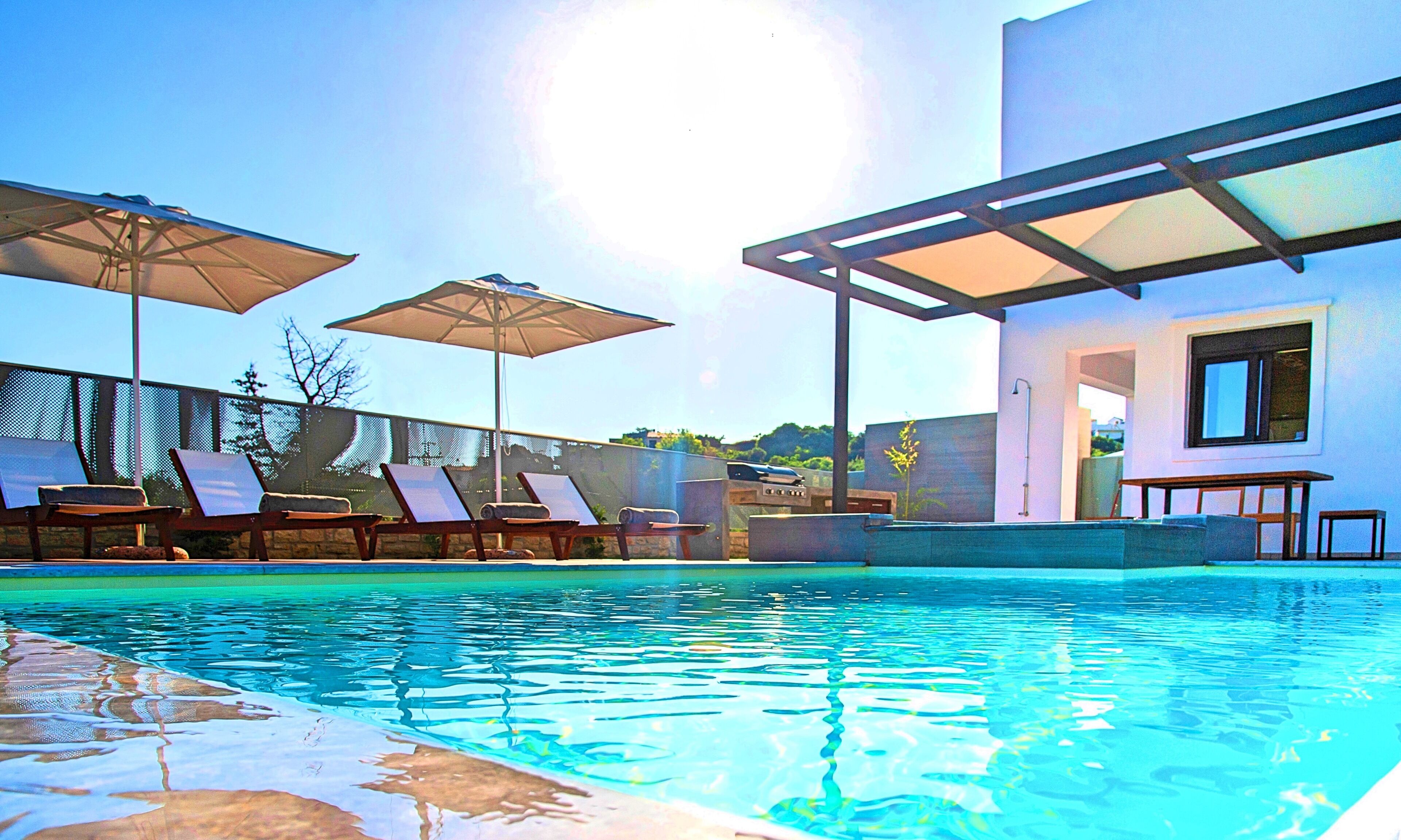 Une piscine avec des chaises longues et des parasols, un bâtiment moderne doté d’une fenêtre, et un ciel dégagé.
