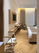 A hotel room with a bed, a desk, a chair, a TV mounted on the wall, and a window with curtains.