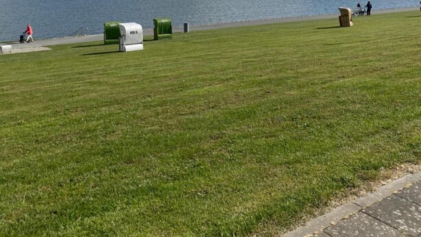 A grassy park area with a body of water, a clear sky, and a few people walking. - Cozy North Sea Apartment – Perfect for You, Your Partner, or Small Family (Büsumer Deichhausen)