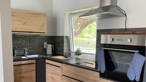 A modern kitchen with wooden cabinets, a built-in oven, and a window with blinds. - Cozy North Sea Apartment – Perfect for You, Your Partner, or Small Family (Büsumer Deichhausen)