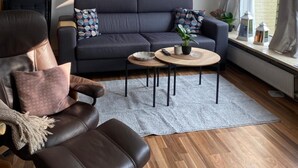 A modern living room with a dark grey sofa, a leather armchair, and a coffee table. - Cozy North Sea Apartment – Perfect for You, Your Partner, or Small Family (Büsumer Deichhausen)