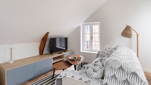 Um quarto com teto inclinado, uma cama com manta estampada, uma escrivaninha com televisão e uma janela com vista.