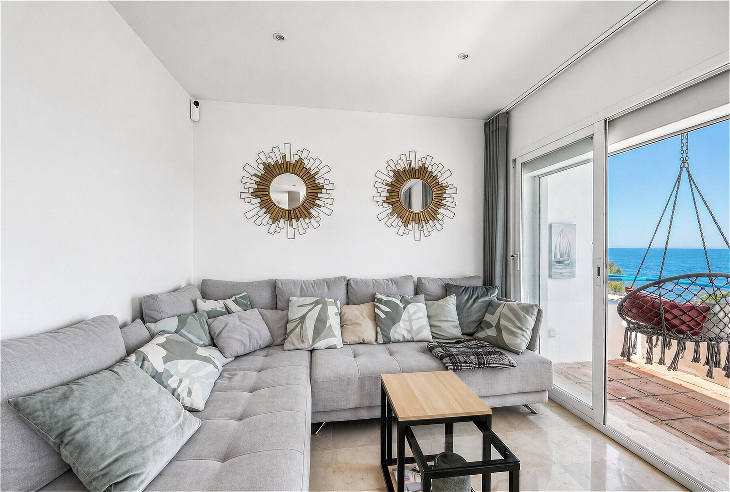 Un salon moderne avec un canapé d’angle gris, deux miroirs muraux, une table basse en bois et une vue sur la mer, visible à travers une porte-fenêtre coulissante.