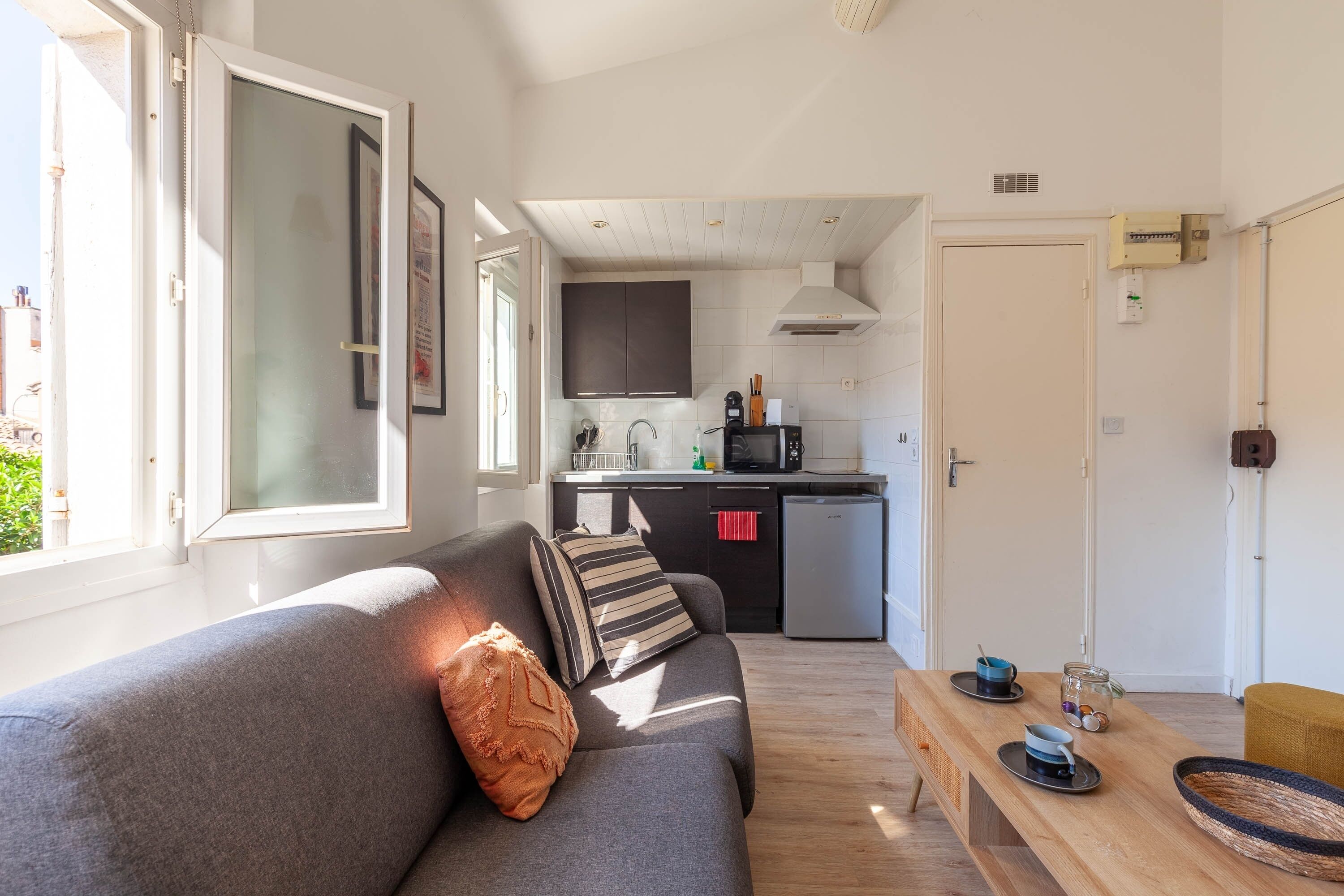 Un salon moderne avec un canapé gris, une table basse en bois et un coin cuisine équipé d’un four à micro-ondes, d’un évier et de placards.