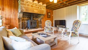 Un salon chaleureux avec une cheminée en pierre, des poutres en bois, un canapé, deux fauteuils à motifs et une petite table.