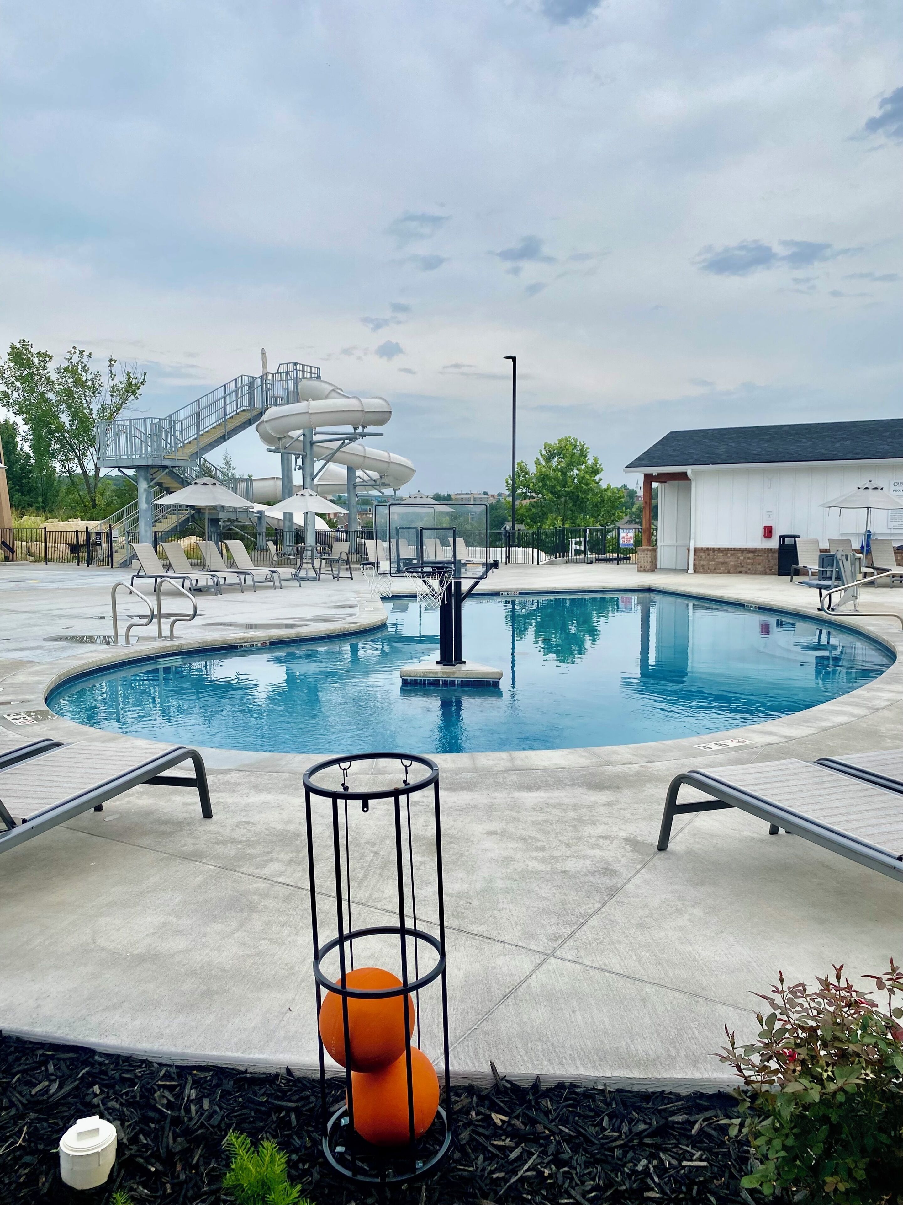 A swimming pool with a water slide, lounge chairs, and a small basketball hoop.
