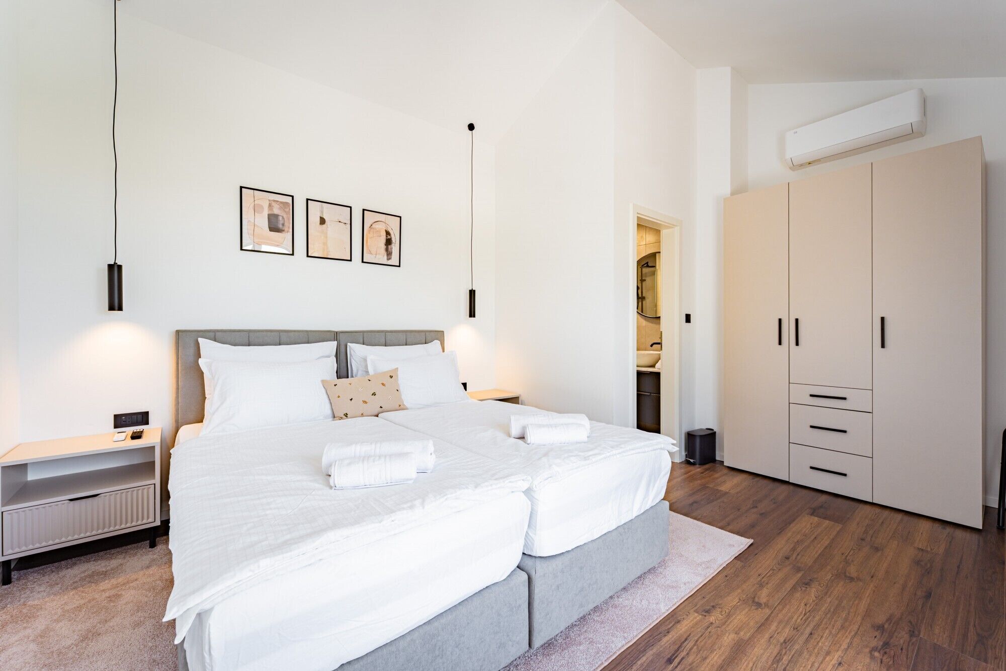 A modern hotel room with a double bed, bedside tables, a wardrobe, and an air conditioning unit.