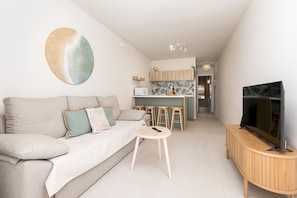 A modern living room with a sofa, a small round table, a TV on a wooden stand, and a kitchen in the background. - Home2Book Chill Terrace & Pool Escape Costa Calma (Costa Calma)