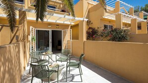 Un patio con mobiliario exterior, una pérgola y un edificio de fachada color beige.