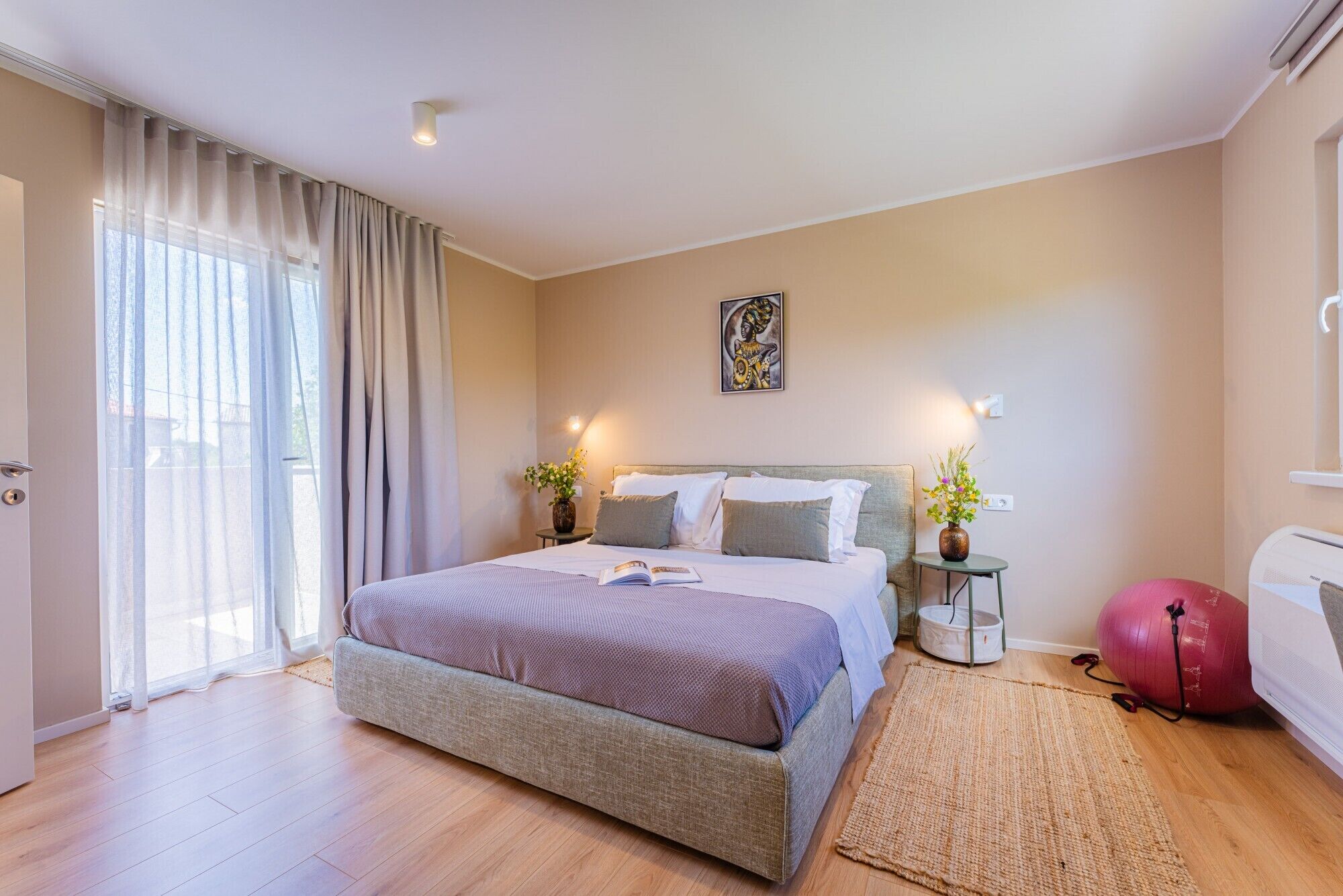 A bedroom with a bed, bedside tables, a wall-mounted artwork, a window with curtains, and a fitness ball.