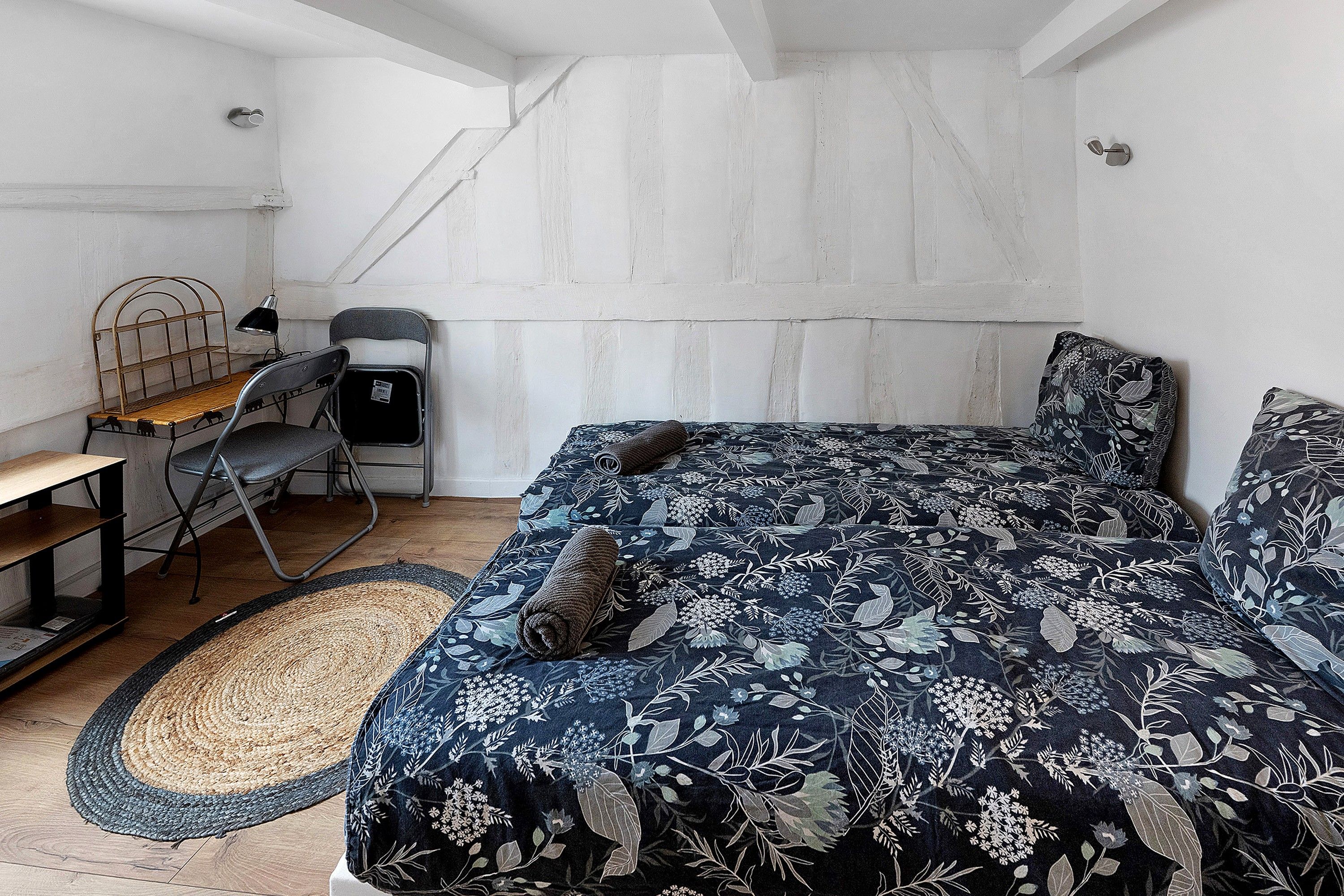 A bedroom with a bed, a desk, a chair, a small stool, and a woven rug.