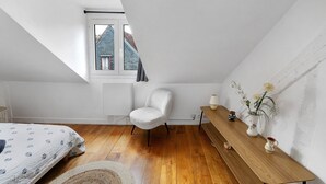 Een slaapkamer met een schuine kap, een enkel raam, een bed met beddengoed met een patroon, een witte stoel, een houten tafel met een vaas en kaarsen, en een kleine plant in een pot.