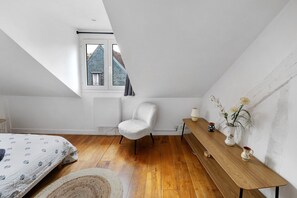 Un dormitorio con techo inclinado, una ventana, una cama con ropa de cama estampada, una silla blanca, una mesa de madera con un florero y velas, y una pequeña planta en maceta.