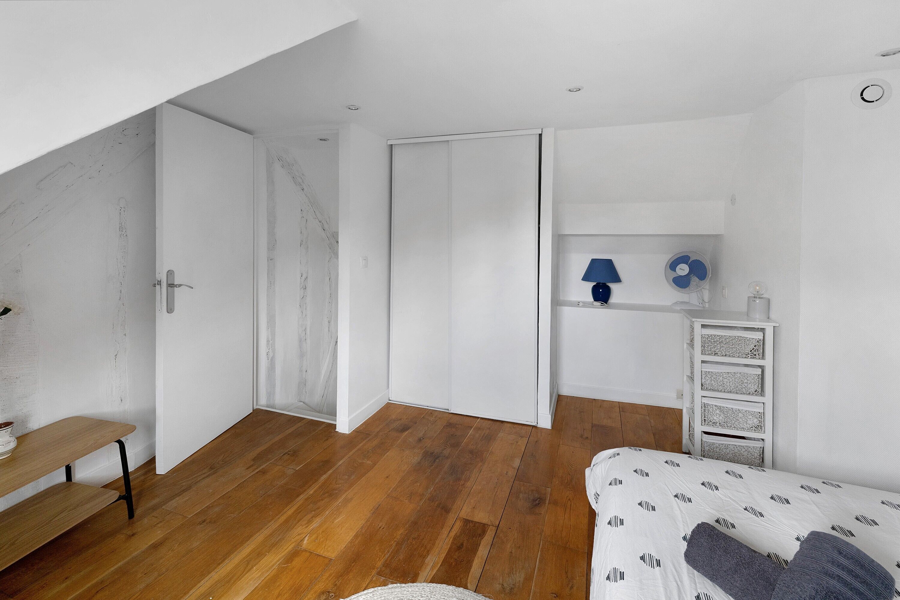 Una camera da letto con pavimento in legno, un letto con copripiumino fantasia, un'armadio bianca, una piccola cassettiera bianca e una lampada blu su uno scaffale.