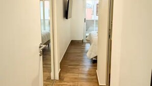 Ein schmaler Flur mit Holzdielen, weißen Wänden und einer Tür zu einem Schlafzimmer mit einem Bett.