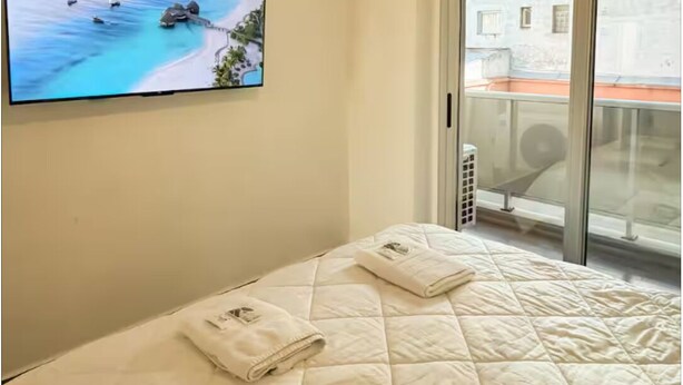 Ein Schlafzimmer mit einem Bett, einem Fernseher, der eine Strandansicht zeigt, einem großen Fenster und einer Deckenleuchte.
