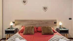 A neatly made bed with red bedding, two nightstands with lamps, and decorative heart-shaped wall art. - Apartment 'Garda Lake' with Shared Pool, Private Terrace, and Air Conditioning (Desenzano del Garda)