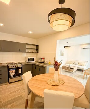 A modern kitchen with a gas stove, microwave, and dark cabinets. There is a dining area with a round table and chairs, and a living room with a sofa and a lamp. - Dpto. a media cuadra de la plaza principal 1 (Salta)