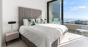 Ein modernes Schlafzimmer mit einem großen Bett, einem Nachttisch, einem transparenten Stuhl und Blick auf die Stadt durch raumhohe Fenster.