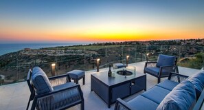 Eine Terrasse mit Sitzgelegenheiten im Freien, einem Glasgeländer und Blick auf den Sonnenuntergang.