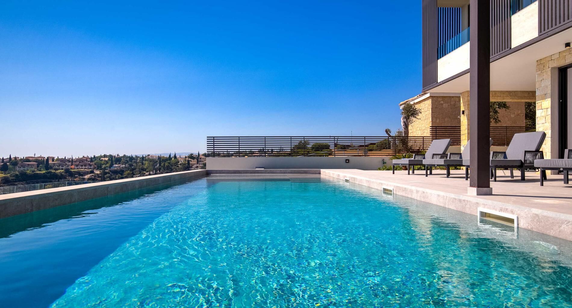 A rooftop infinity pool with clear blue water, a modern building with balconies, and a view of a distant town.