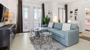 Ein modernes Wohnzimmer mit einer Couch, zwei Beistelltischen, einem Fernseher und einem Balkon mit Vorhängen.