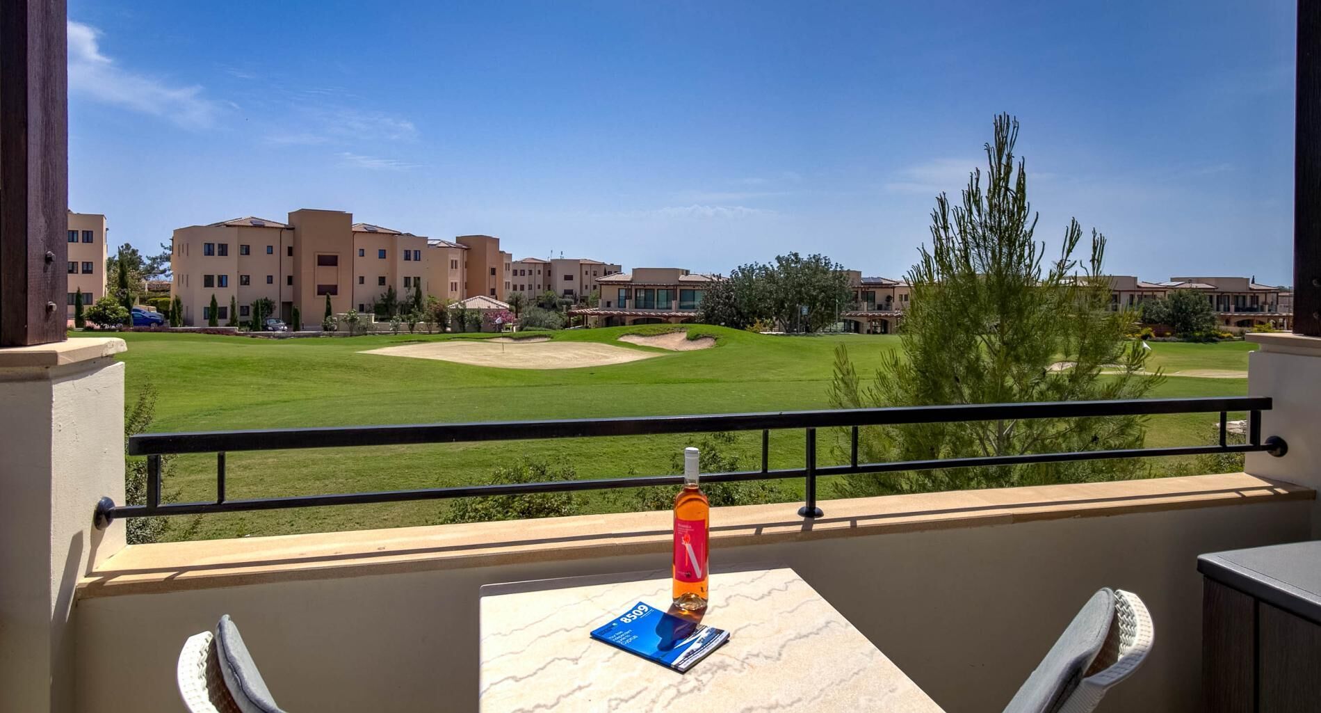 A balcony with a view of a golf course, a bottle, and a menu on a table.