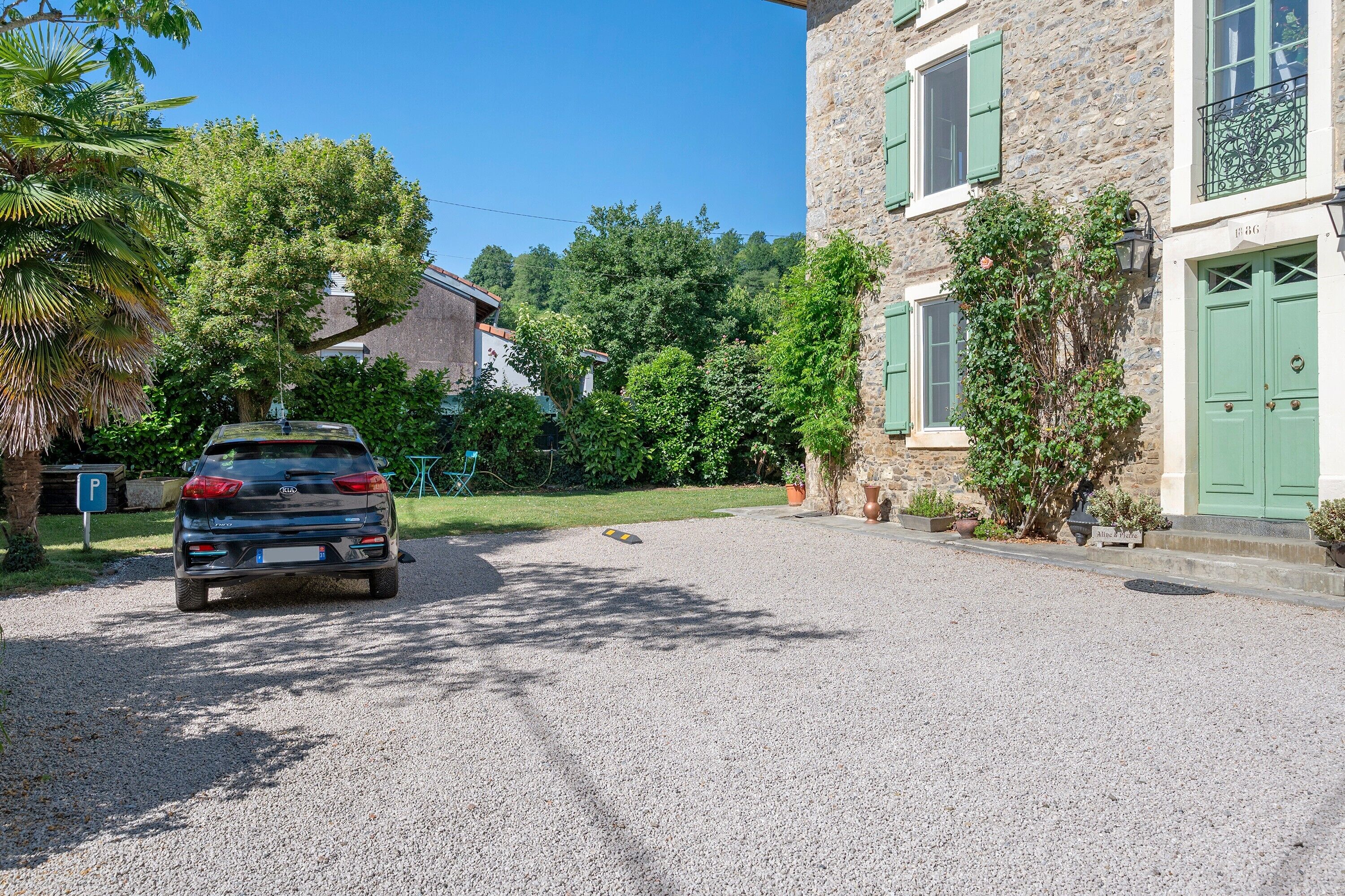 A car parked in front of a stone building with green shutters, a gravel driveway, and lush greenery.