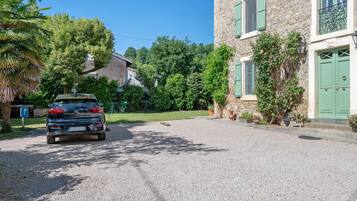 A car parked in front of a stone building with green shutters, a gravel driveway, and lush greenery.
