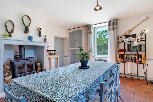 A room with a fireplace, a table with a patterned tablecloth, chairs, a potted plant, and a shelf with bottles.