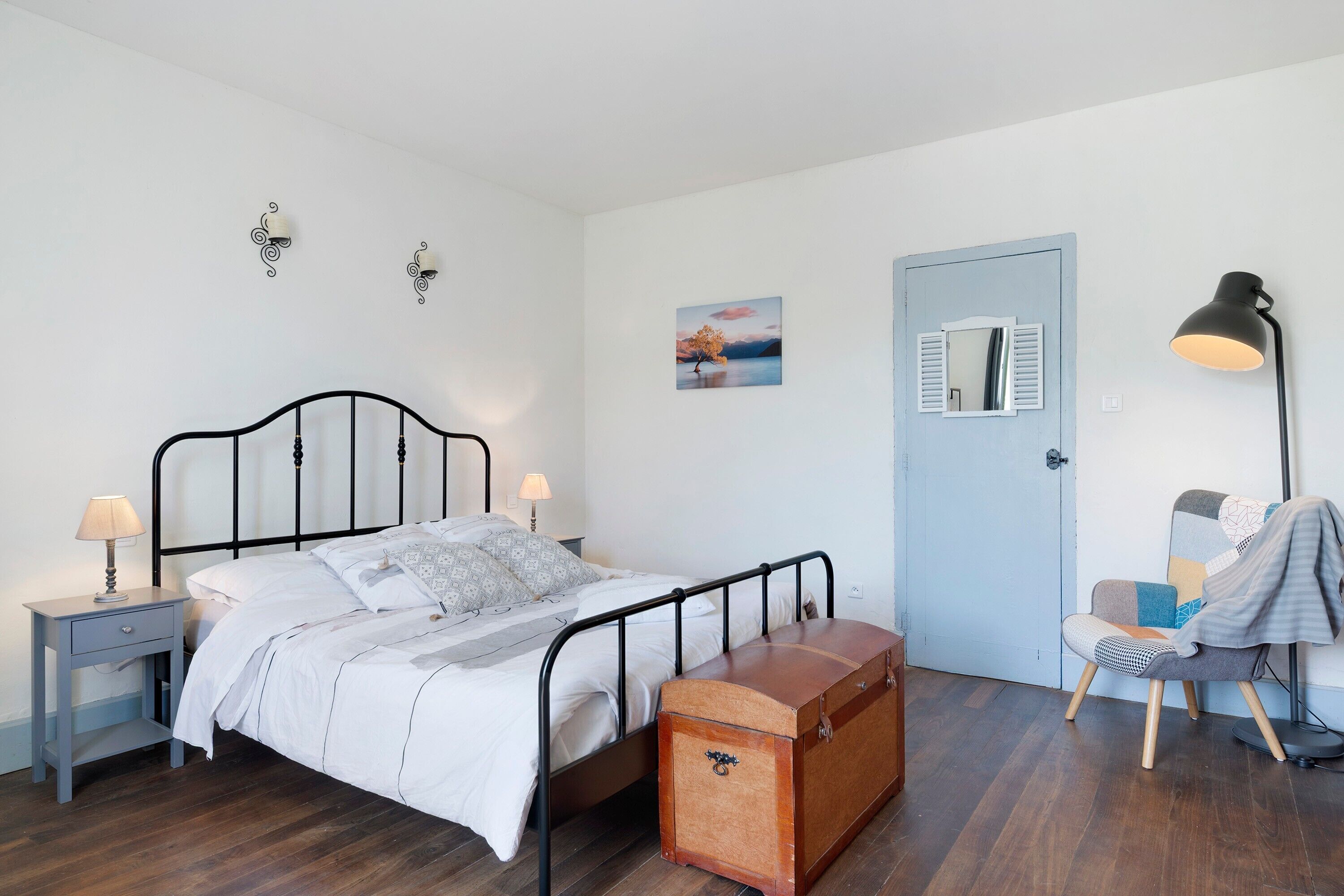 Una camera da letto con un letto, comodino, lampada, cassettiera, sedia e una porta.