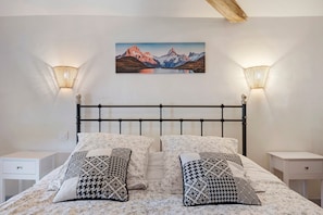 A bedroom with a bed, two bedside tables, a wall-mounted lamp, and a painting of mountains.