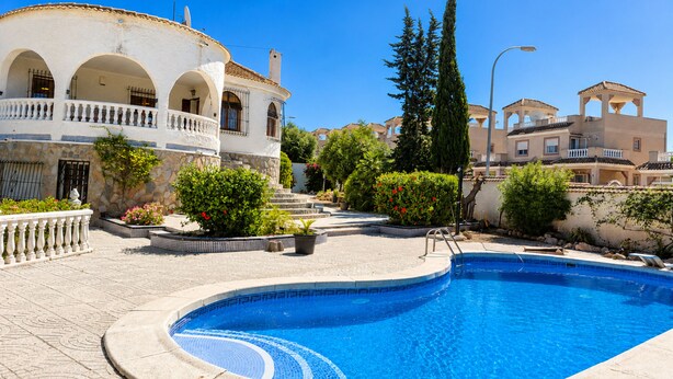 A swimming pool with a tiled edge, surrounded by a stone-paved area, and adjacent to a building with arches and balconies. - Private Pool Villa La Siesta (Torrevieja)