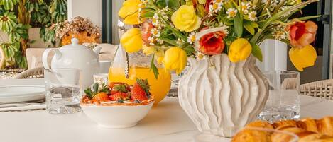 A breakfast table with a vase of flowers, a bowl of fruit, a teapot, and croissants.