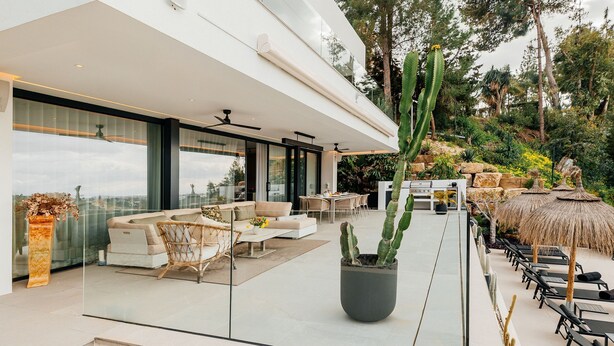 A modern outdoor living area with a glass wall, a large potted cactus, outdoor furniture, and a dining area. - Modern Luxury Villa with Panoramic  View (Marbella)