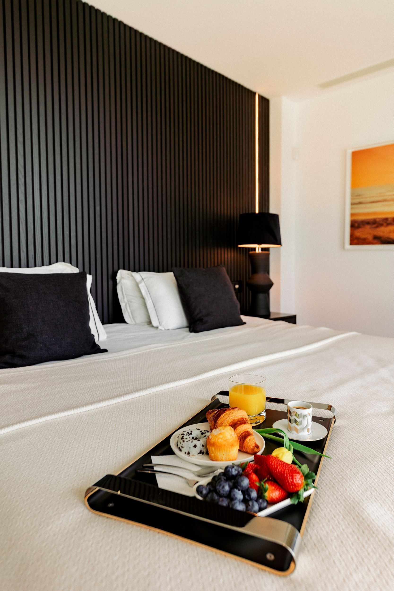 Cama arrumada com uma bandeja de itens de café da manhã, incluindo croissants, frutas e bebidas, em quarto de hotel moderno com painel de madeira escura e um quadro na parede.