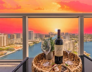 A bottle of wine, two wine glasses, and a woven basket on a balcony with a cityscape view. - Top-Floor Waterway 2BR · Beachwalk Resort FL (Hallandale Beach)