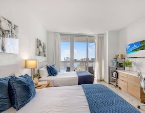 A modern hotel room with a bed, bedside table, lamp, and a view of the city through large windows. - Top-Floor Waterway 2BR · Beachwalk Resort FL (Hallandale Beach)