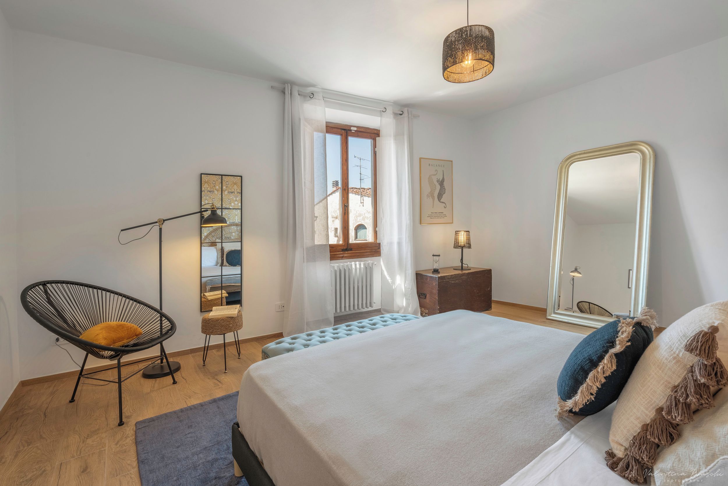 A bedroom with a bed, a chair, a bedside table, a mirror, and a window with curtains.