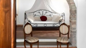 Een kamer met een wit slaapbank onder een hemelbed, twee gestoffeerde fauteuils en een bakstenen muur.