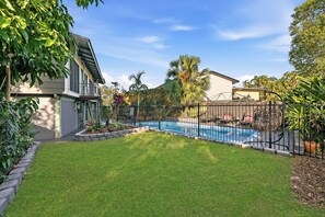 A residential backyard with a swimming pool, surrounded by a fence, and featuring a well-maintained lawn and various plants.