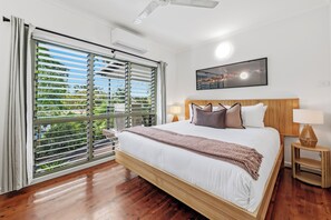 A hotel room with a large bed, a window with blinds, a ceiling fan, a wall-mounted picture, and a bedside table with a lamp. - Tropica Lookout A Serene Coastal Pool House (Rapid Creek)