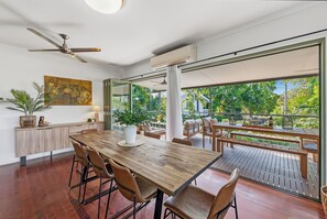 A modern dining area with a wooden table, chairs, and a view of a lush garden through large windows. - Tropica Lookout A Serene Coastal Pool House (Rapid Creek)