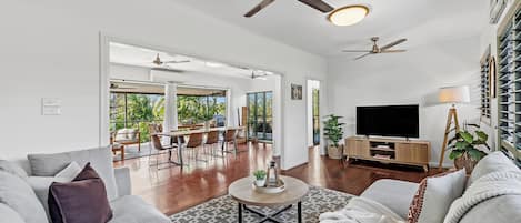 A spacious living room with a large sofa, a coffee table, and a TV stand. There are ceiling fans and a dining area visible in the background.