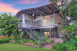 A modern two-story house with a covered balcony, a staircase, and lush greenery surrounding it.