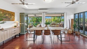 Un vaste espace à manger avec une table et des chaises en bois, de grandes fenêtres donnant sur un paysage verdoyant, des ventilateurs de plafond et une cuisine moderne dotée d’un plan de travail en bois.