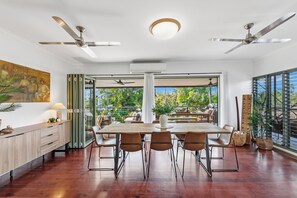 A spacious dining area with a wooden table and chairs, large windows with a view of greenery, ceiling fans, and a modern kitchen with a wooden countertop.