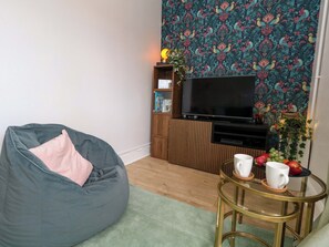 Ein Wohnzimmer mit Sitzsack, Flachbildfernseher, Bücherregal und einem kleinen Tisch mit Tassen und Obst.