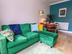 A green sofa with a blue throw, a gold floor lamp, a dining table with chairs, and a radiator. - Seagull and Chips (Weymouth)
